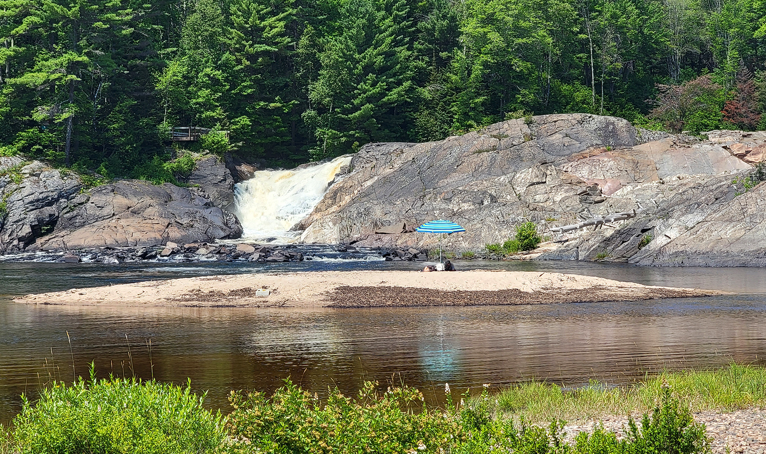 a person under a beach umbrella relaxes on a large bar of soft tawny sand surrounded by smooth rocky banks and thick green boreal forest on a summer day.