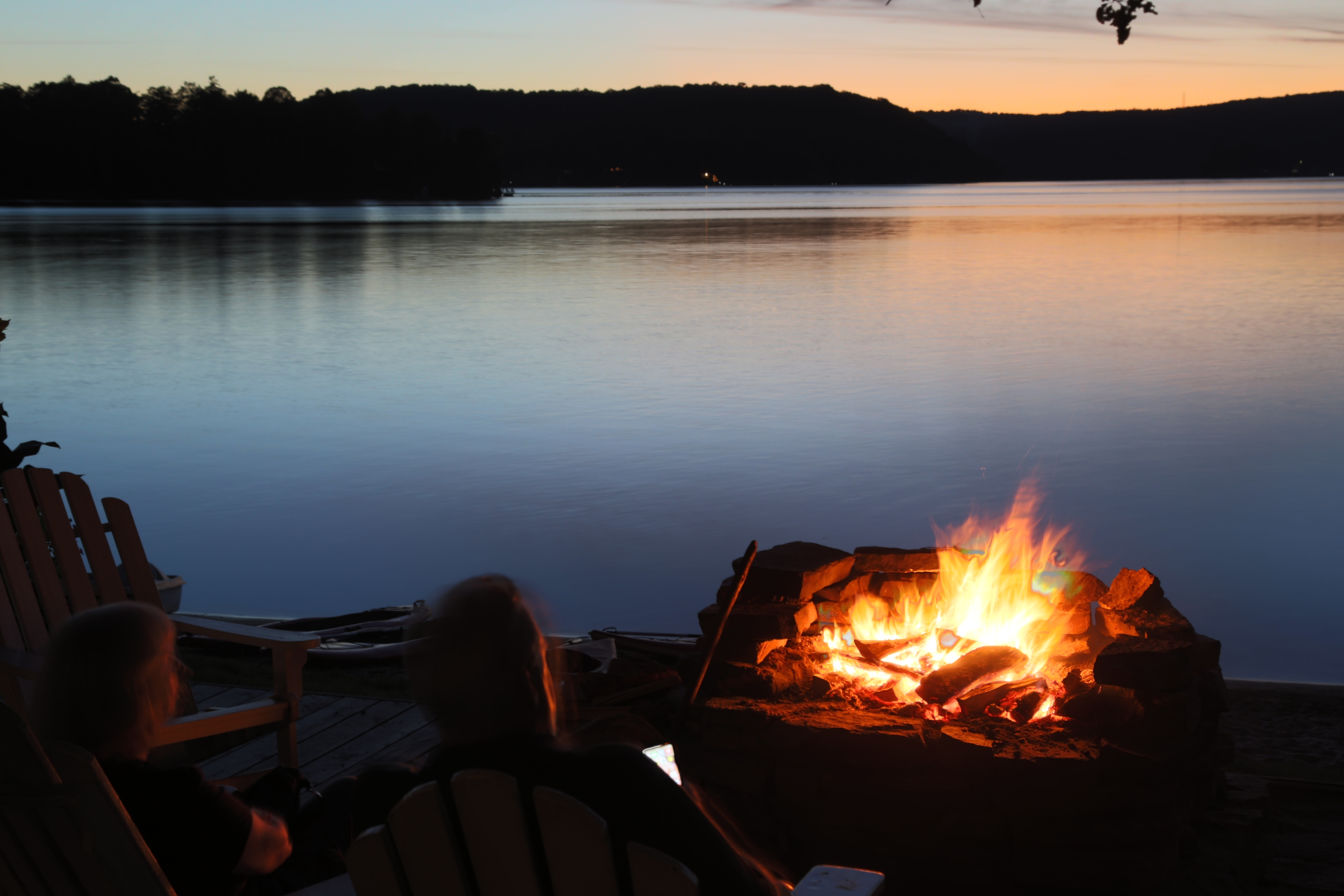 a crackling orange campfire glows in the twilight at the edge of a glassy lake reflecting the last of a clear orange sunset. 