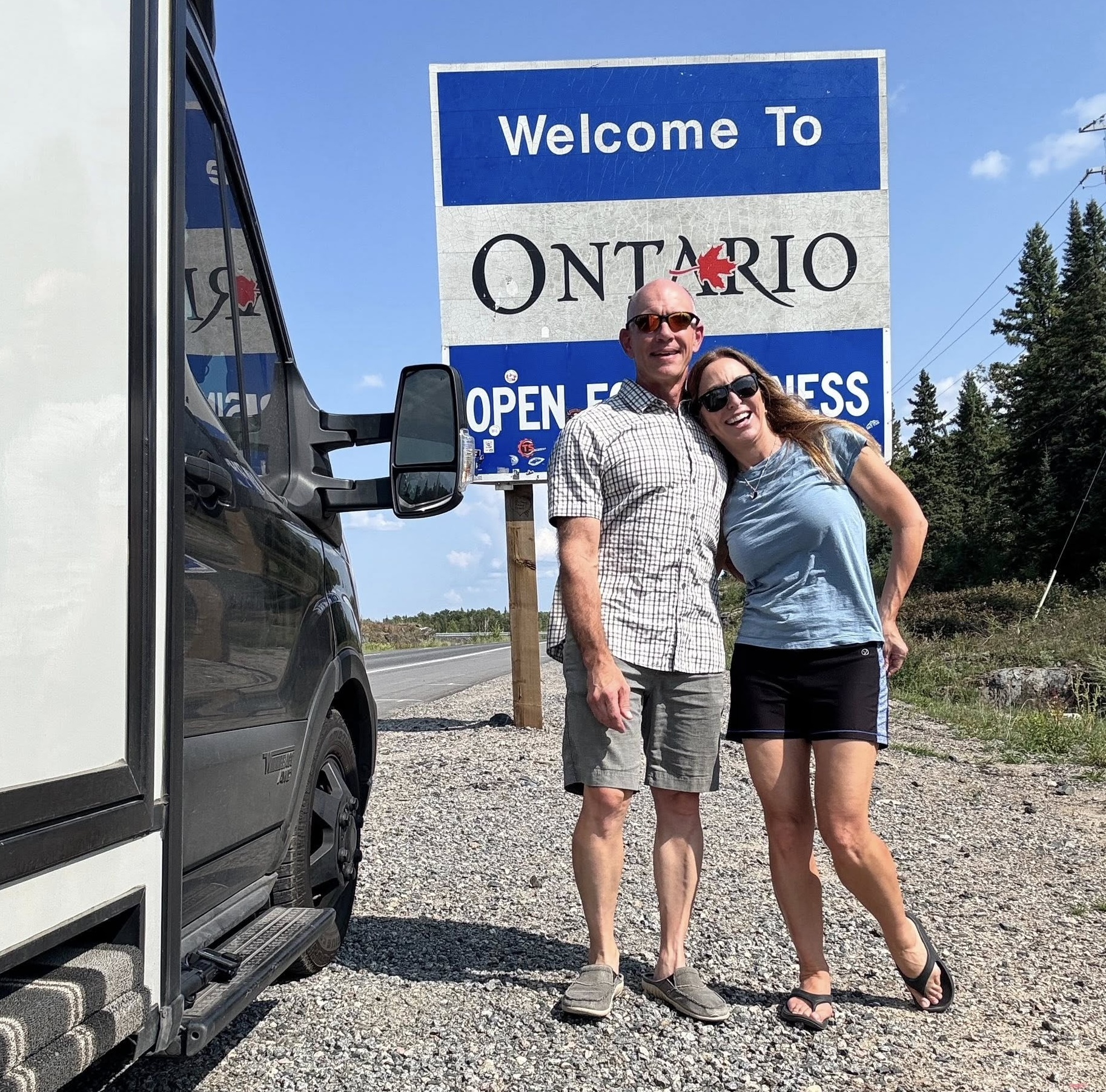 Stef and James of The Fit RV smile with their arms around each other's shoulders, standing next to their RV at the roadside in front of a sign that says "Welcome to Ontario", under a bright blue sky. 