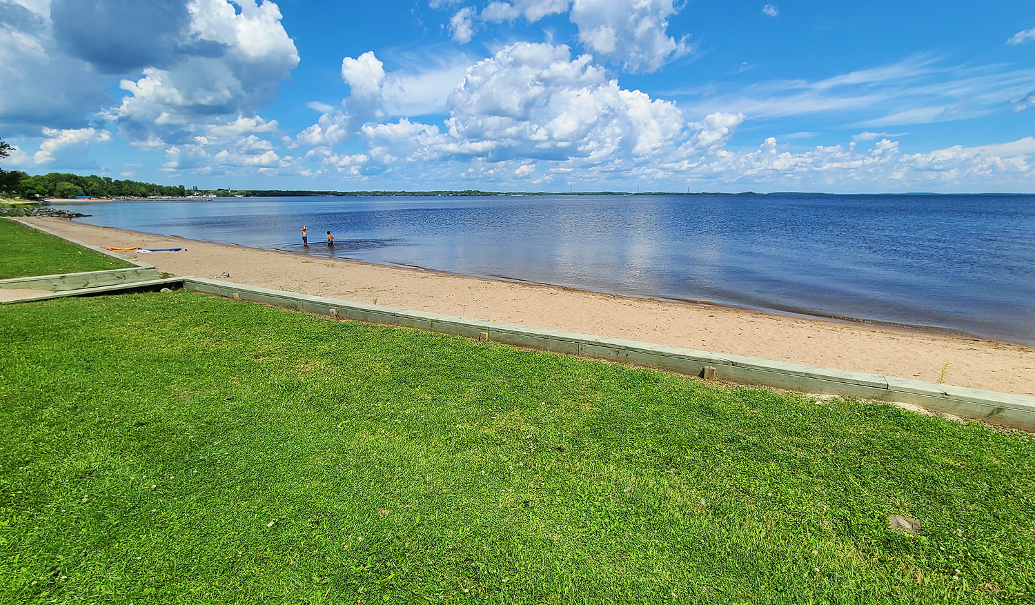 a white sandy beach edge in emerald green lawn runs the perimeter of a glassy blue lake under a bright blue summer sky.