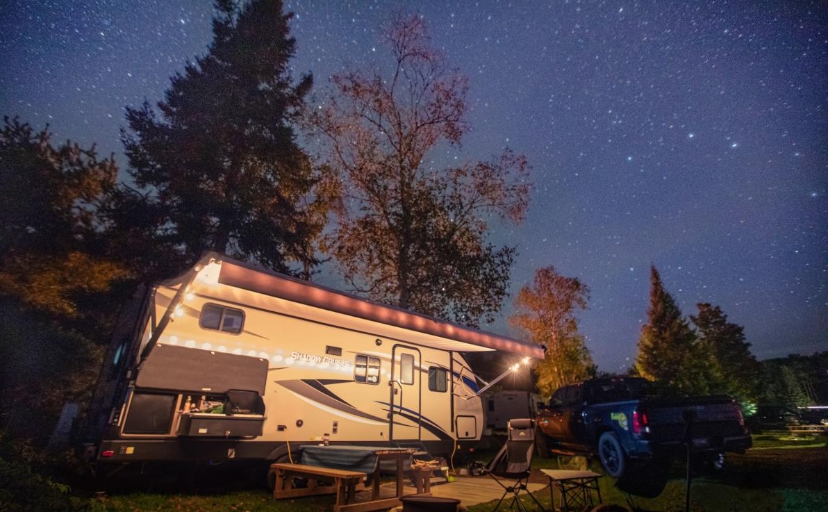 An RV lit up in the night next to leafy trees under a clear starry sky.