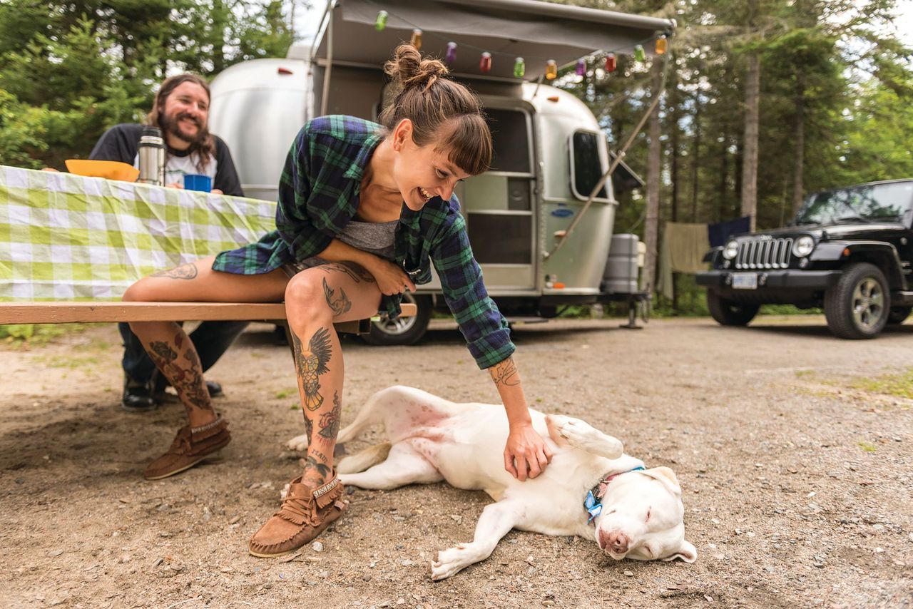 a smiling woman sitting at a campsite picnic table with her partner scratches the belly of a dog laying on the ground next to her. Their RV and a forest is in the background.