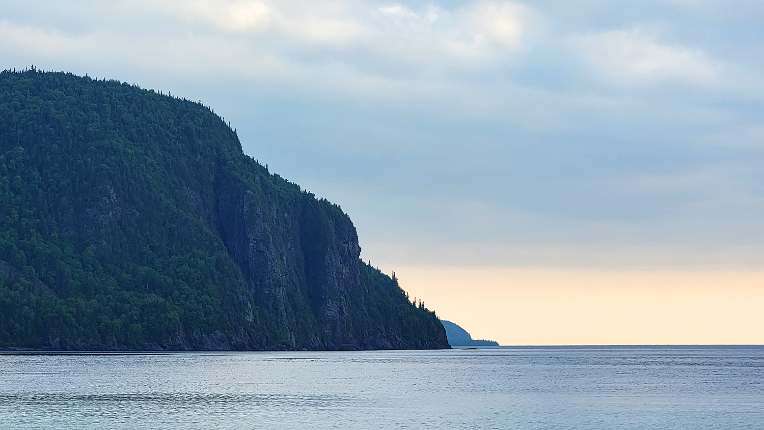 The tall, dark rock formations of Old Woman Bay, sloping down to the silvery Lake Superior water under soft sunset clouds of cool blue and gold.