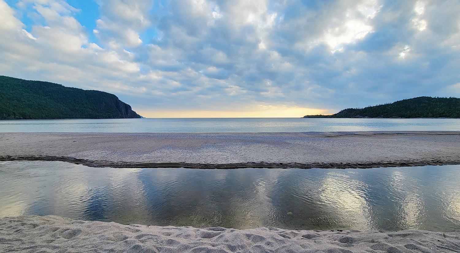 flat bars of soft sand around shallow pools of glassy water in front of Lake Superior and Old Woman Bay. Dark sloping rock formations sit on the horizon under a summer evening sky.