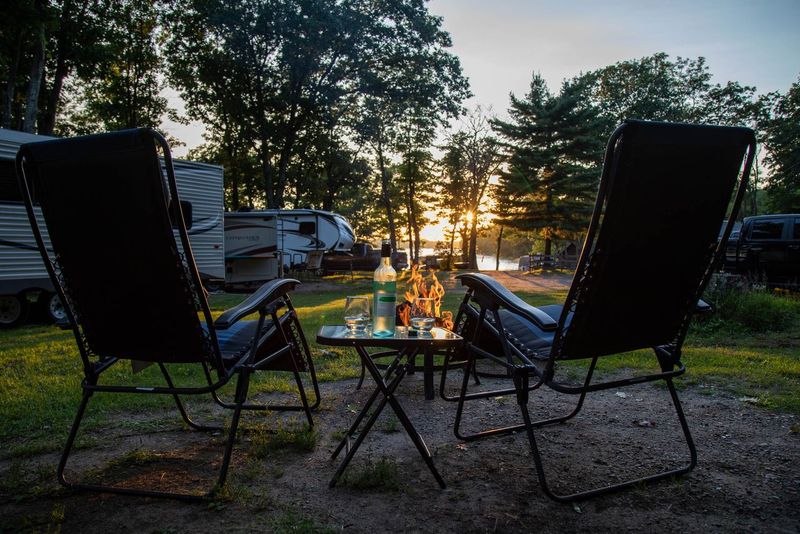 two chairs and a table set side by side facing a campfire and a setting sun at a forested RV park.