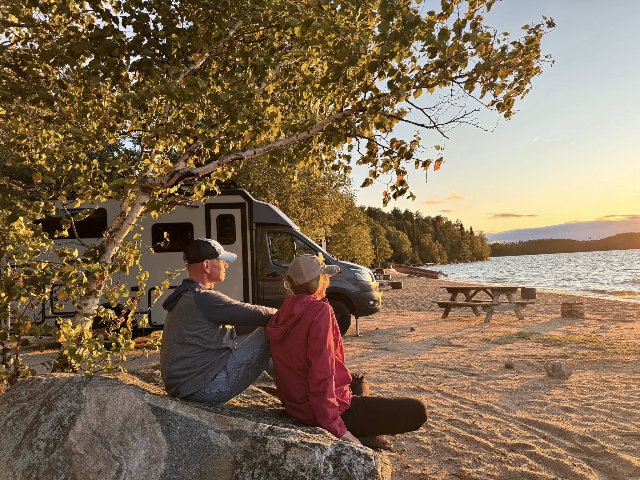 A man and woman sit on a large rock on a sandy beach next to green forest, looking out over a calm lake at the sun sets. 