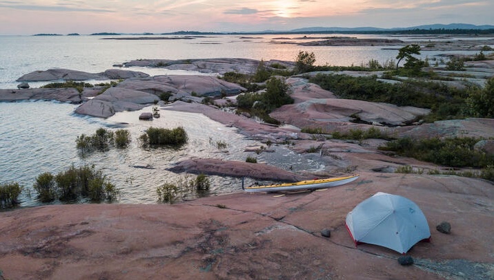 a small tent sits on a smooth rock shore dotted with green shrubbery, overlooking a vast lake lit by a soft pink sunset.
