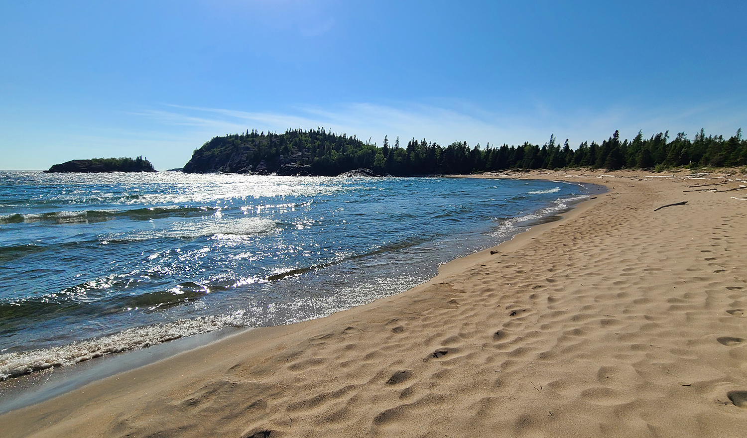 a wide swatch of fluffy white sand beach skirts shimmering Lake Superior waves on a clear summer day.
