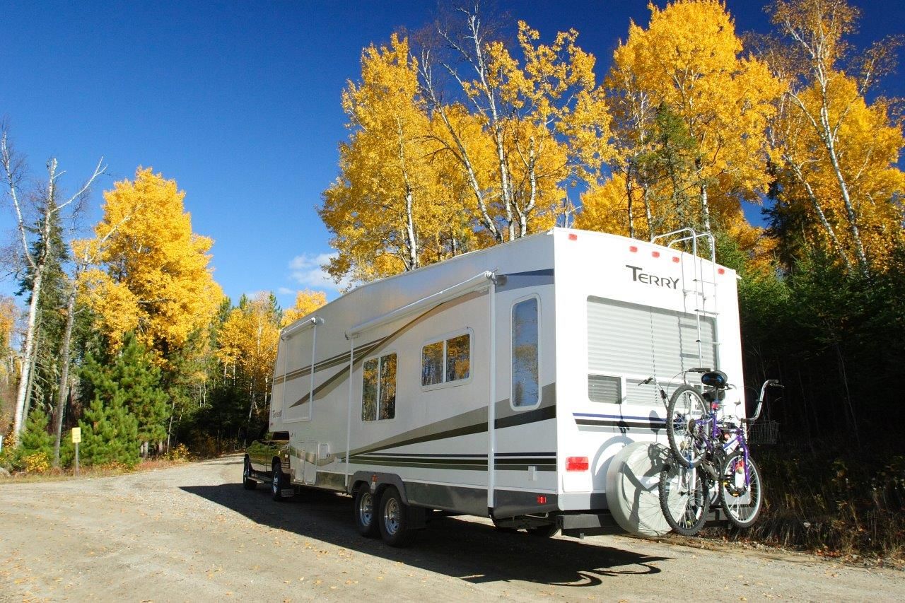 An RV drives down an empty road through bright yellow fall forest under a vivid, clear blue sky.