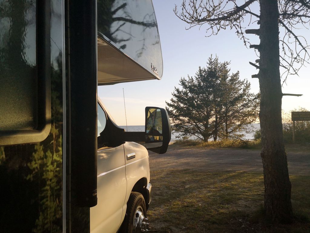 An RV parked facing a broad lake view lit by a golden, misty sunrise in the summer.