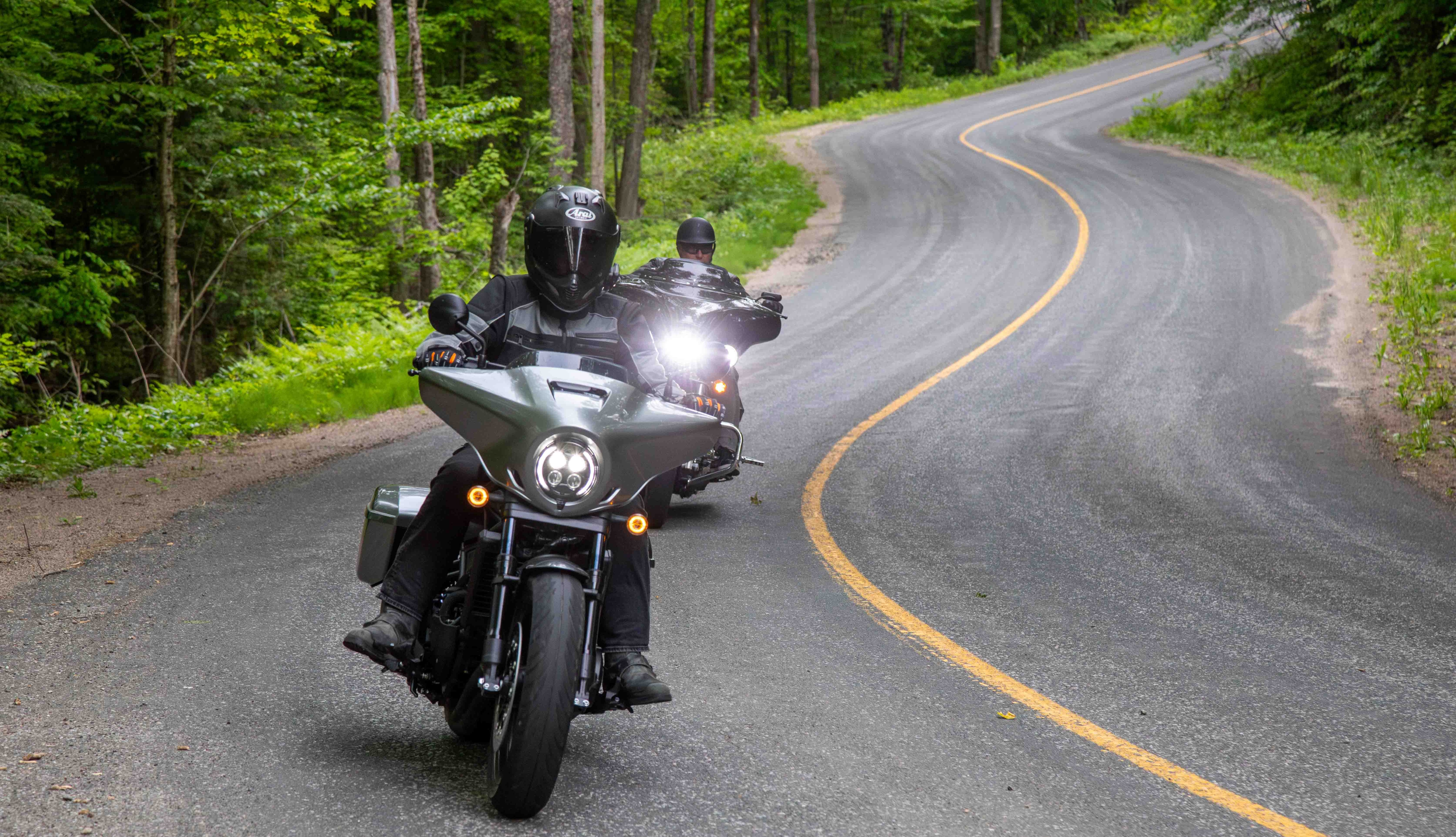 Two motorcyclists ride down a widing paved paved road through lush green forest.