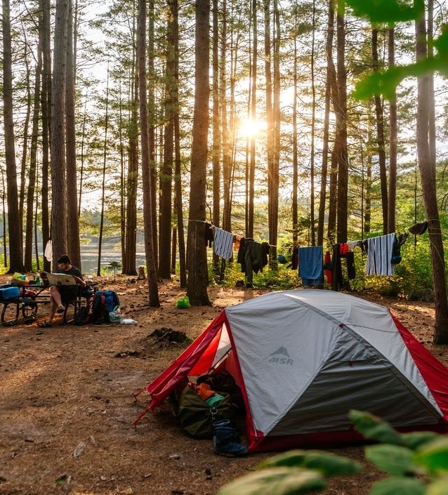 a camper relaxes at their campsite as a setting sun peeks through tall pine trees.
