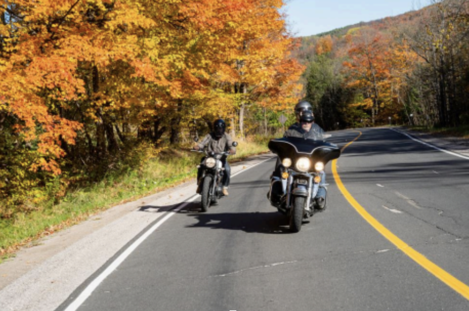 Two motorcyclists ride down a winding paved road through blazing orange autumnal trees on a bright sunny fall day. 