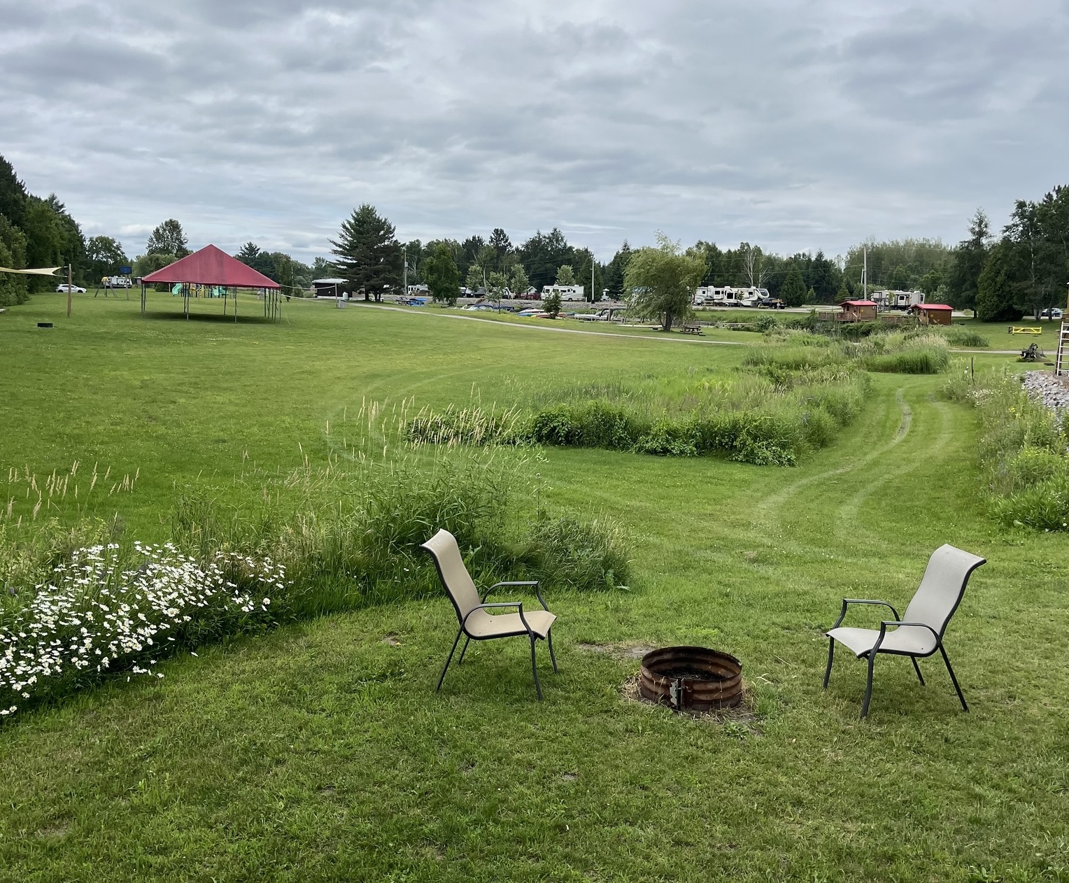 two chairs sit facing a fire pit in a lush green field with a path winding up to and RV park and forest.