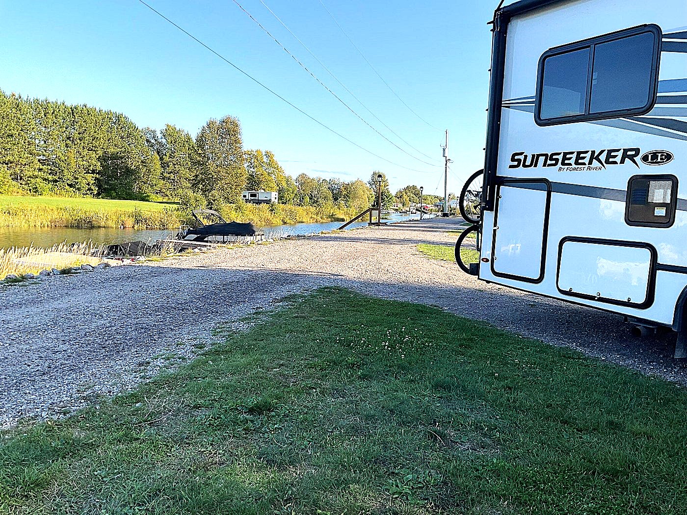 an RV parked at a grassy site next to a river with lush forested banks under a clear blue sky.