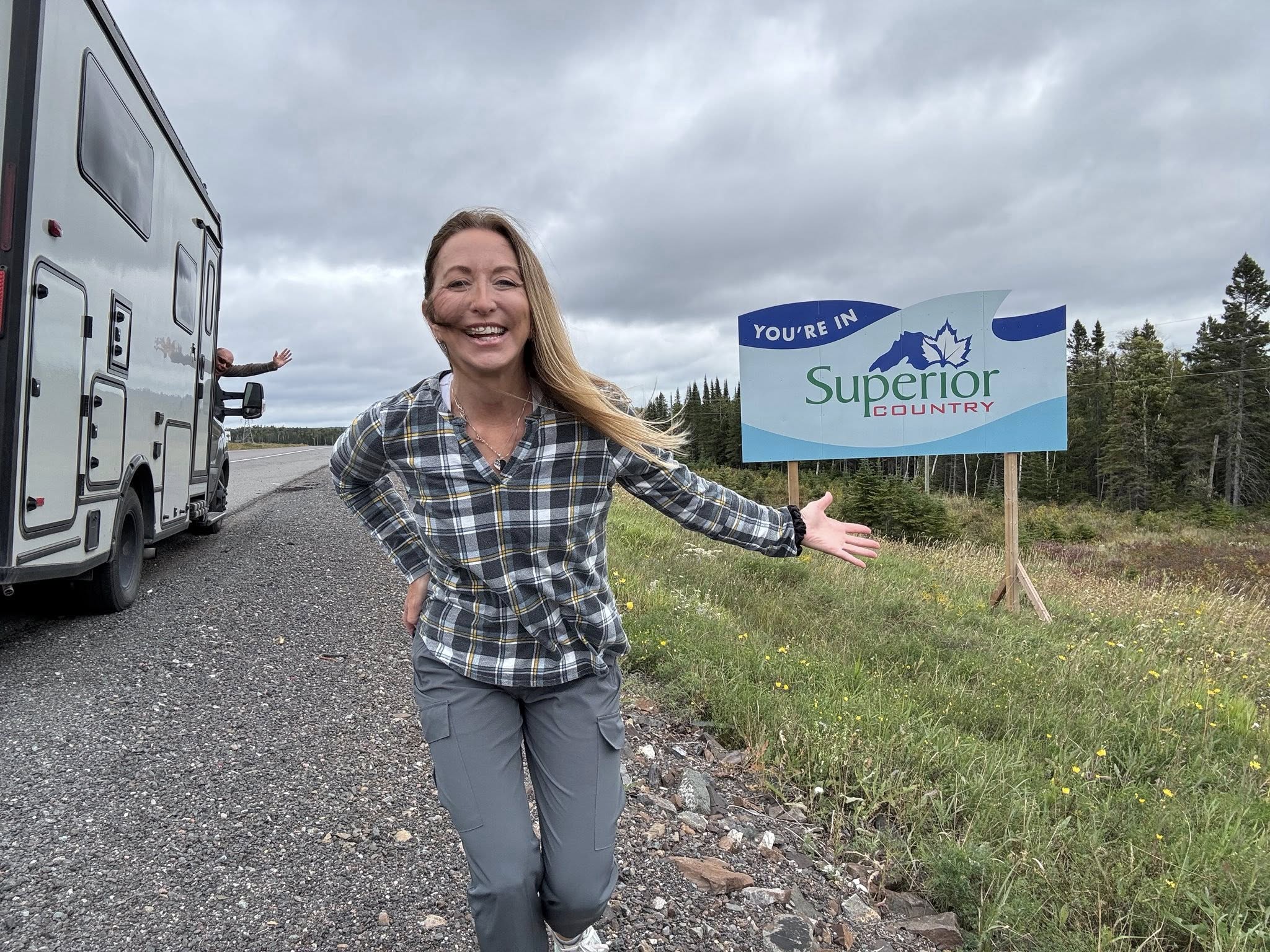 a smiling Stef from The Fit RV gestures to a roadside sign behind her welcoming visitors to Superior Country.
