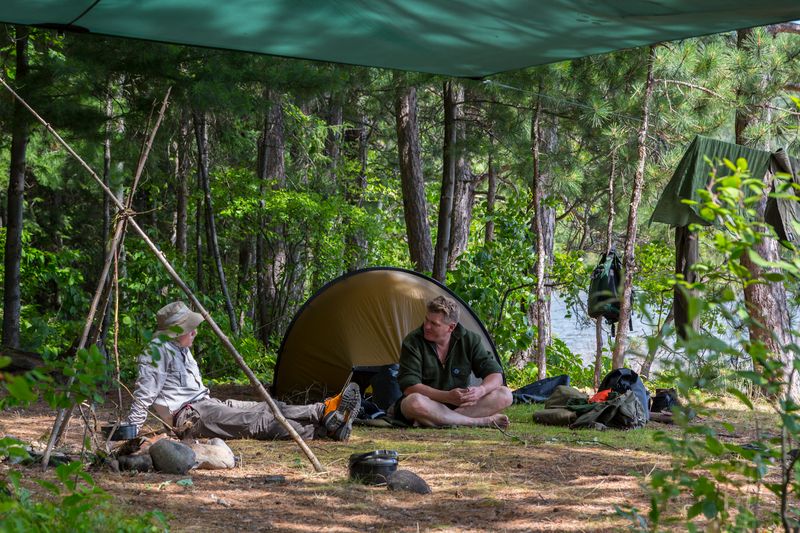 two people relax under a tarp shelter supported by long natural sticks next to their tent, happily chatting deep in a shady green summer forest. A lake peers through the trees in the distance.