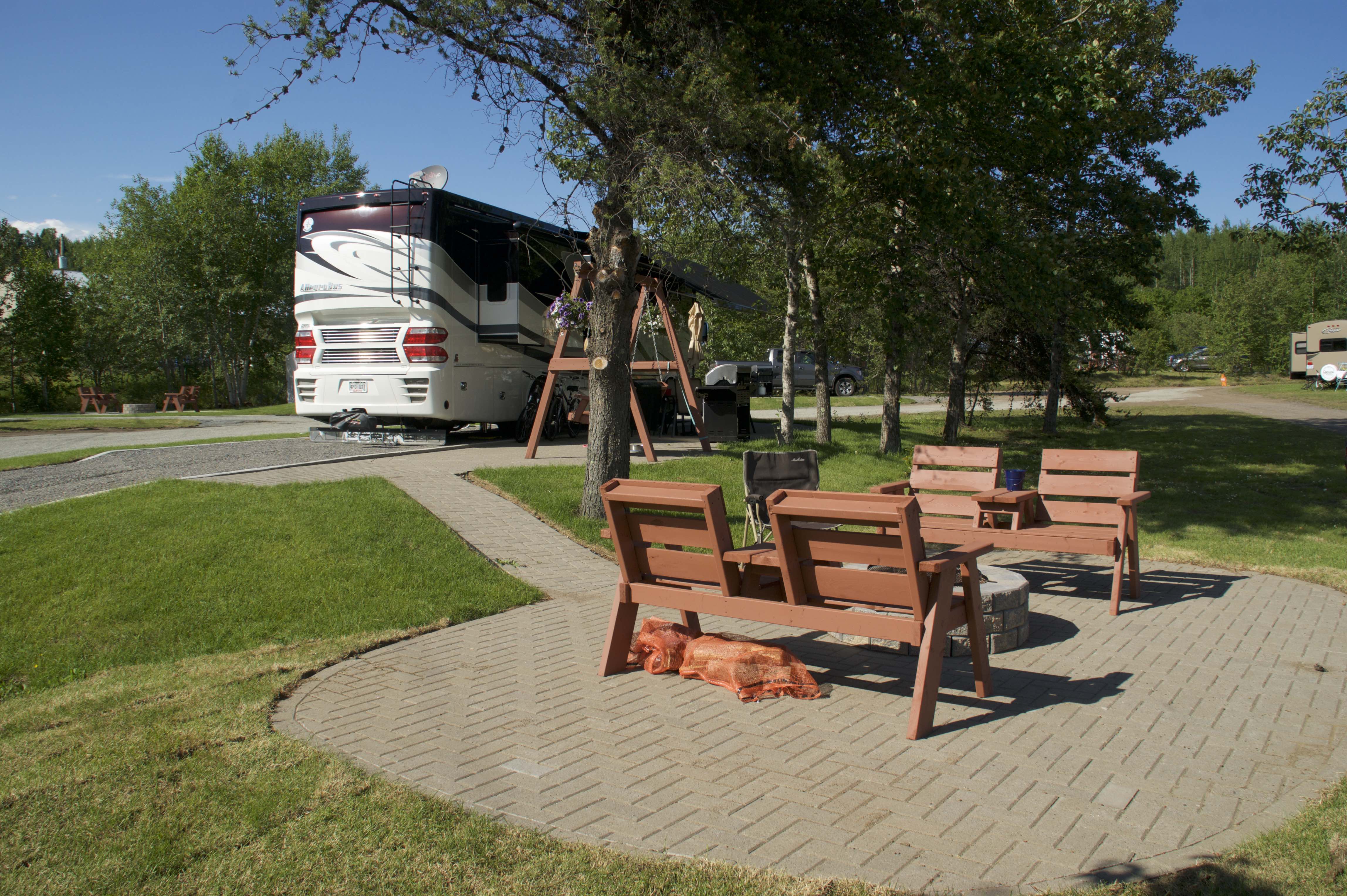 An RV parked next to a paved fire pit area with wooden lounge chairs on a summer day. 
