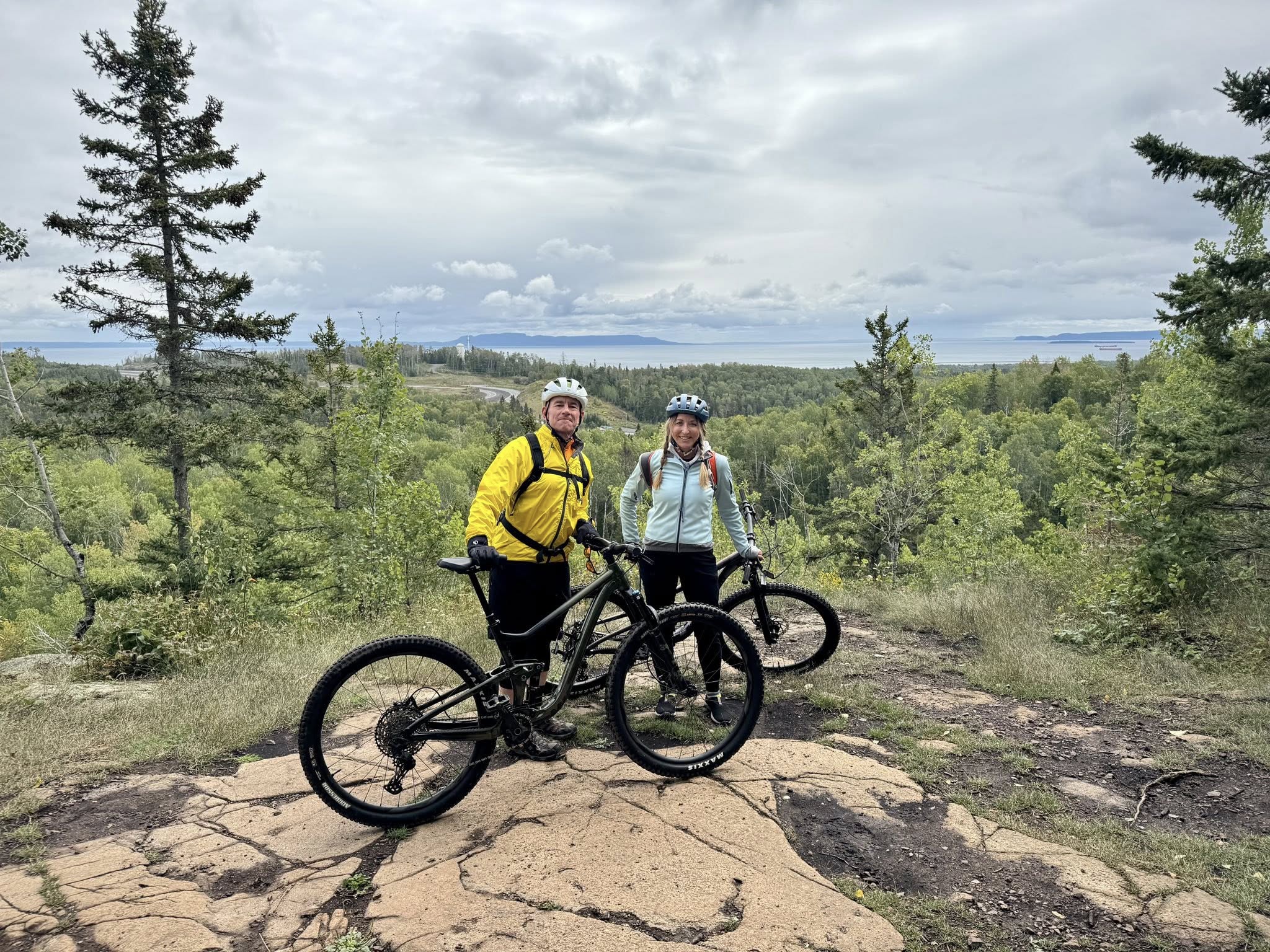 A smiling man and woman stand next to their bikes at a rocky forested view over Lake Superior on the Trowbridge trail.