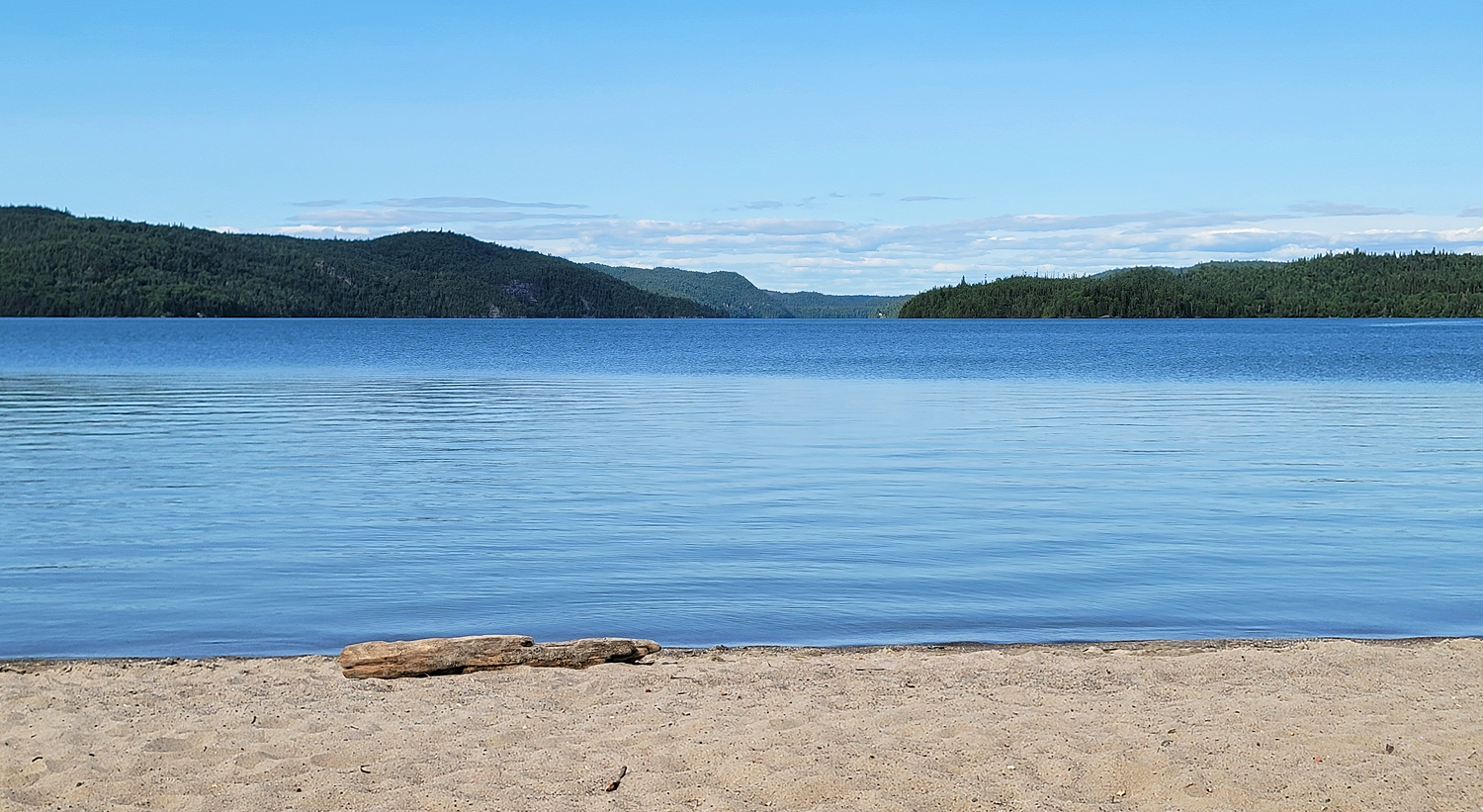 A soft sand beach meets clear blue water and sky intersecting with rugged forested cliffs on the horizon.