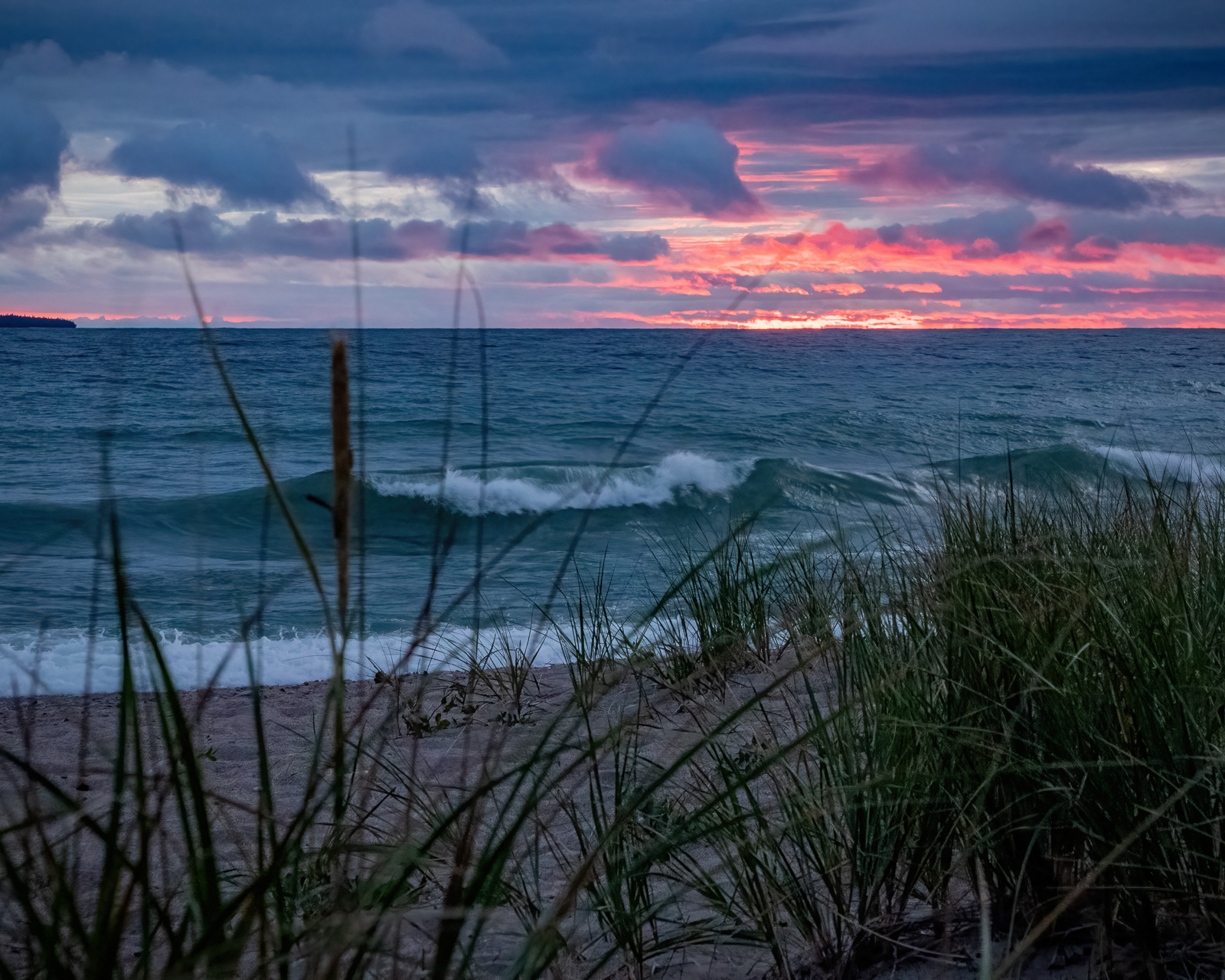 a deep purple and pink sunset over the moody grey waves of Lake Superior. Grasses and shrubbery on the gravelly bank rustle in the breeze.