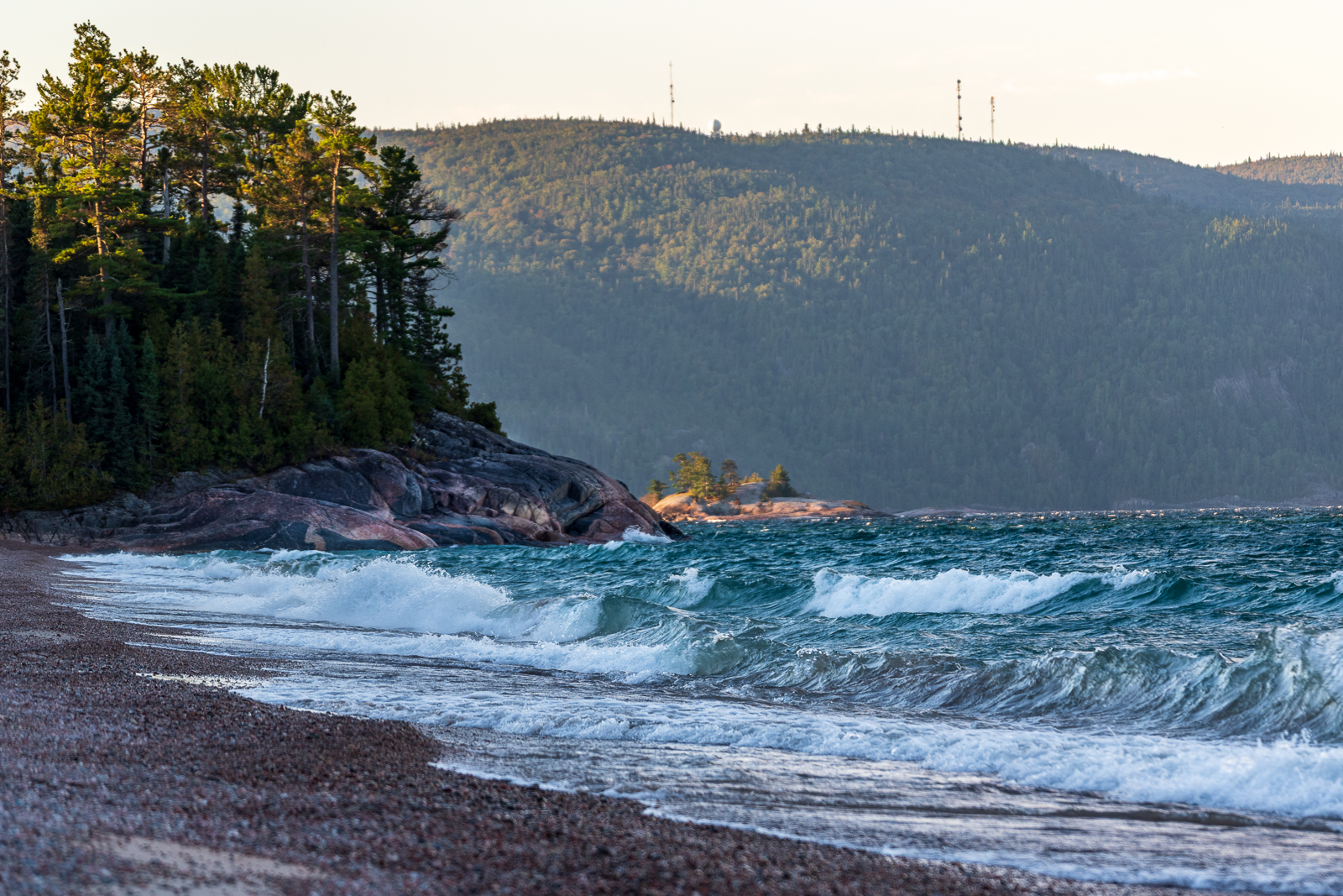 light blue waves crash against a sandy beach next to a rock cliff covered in boreal forest.