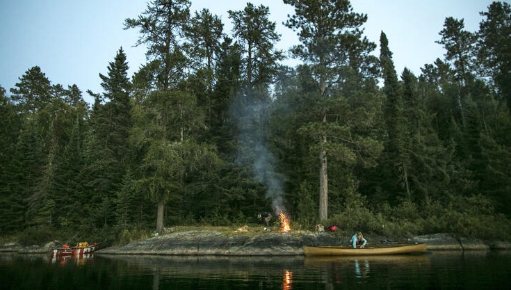 a person looks through the supplies in a canoe at the water's edge next to a campsite on a small island surrounded in thick green boreal forest under a clear summer sky.