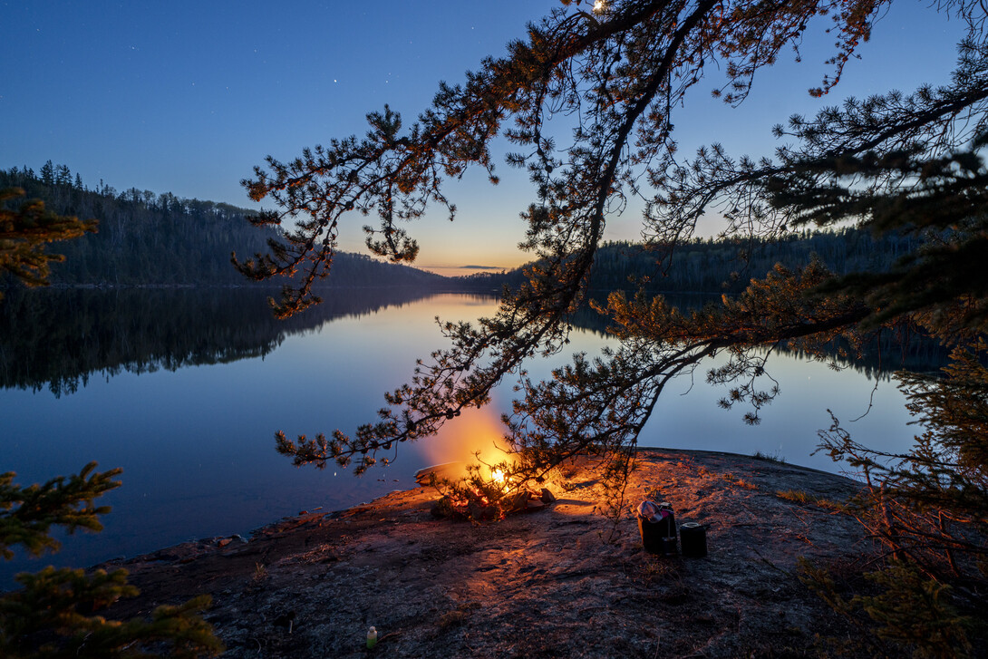 a glowing orange campfire lights up a smooth rock bank overlooking a glassy lake perfectly reflecting a darkening evening sky. Pine bows stretch out overhead.
