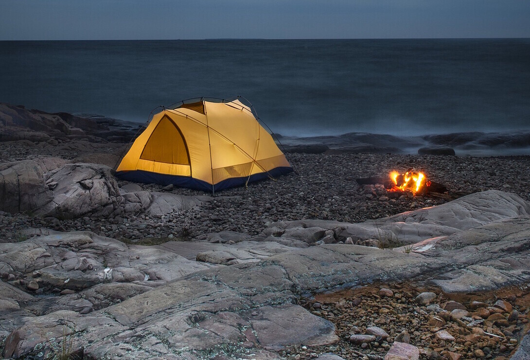 a glowing tent sits next to a campfire on a rocky shore next to the misty splashing waves of Lake Superior, looking grey in the darkening evening.