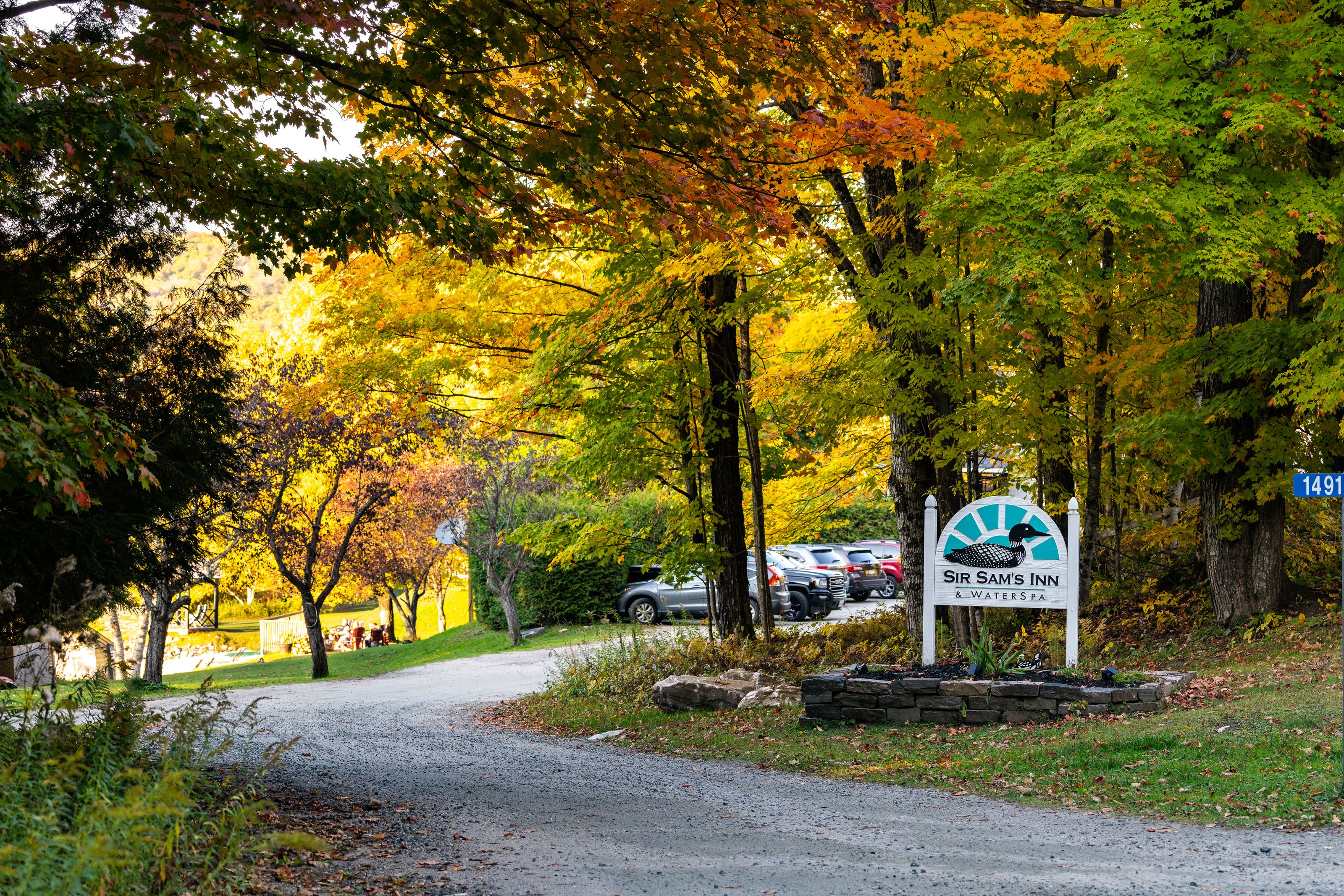 a gravel lane winds through bright autumn trees in yellow, red, green and gold. A wooden sign at the edge of the road welcomes visitors to Sir Sam's Inn & Spa. 