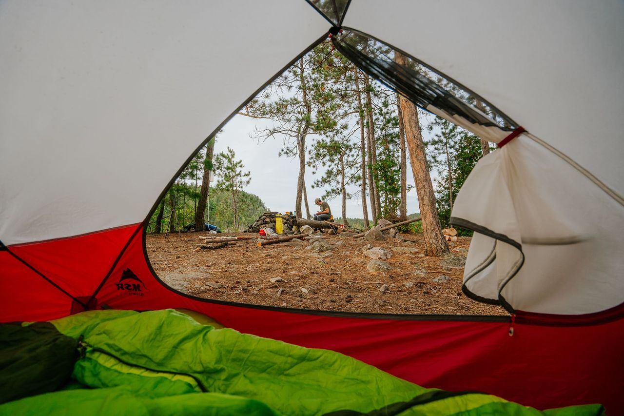 an open tent flap, showing a person relaxing on a cliff's edge in a pine forest outside.