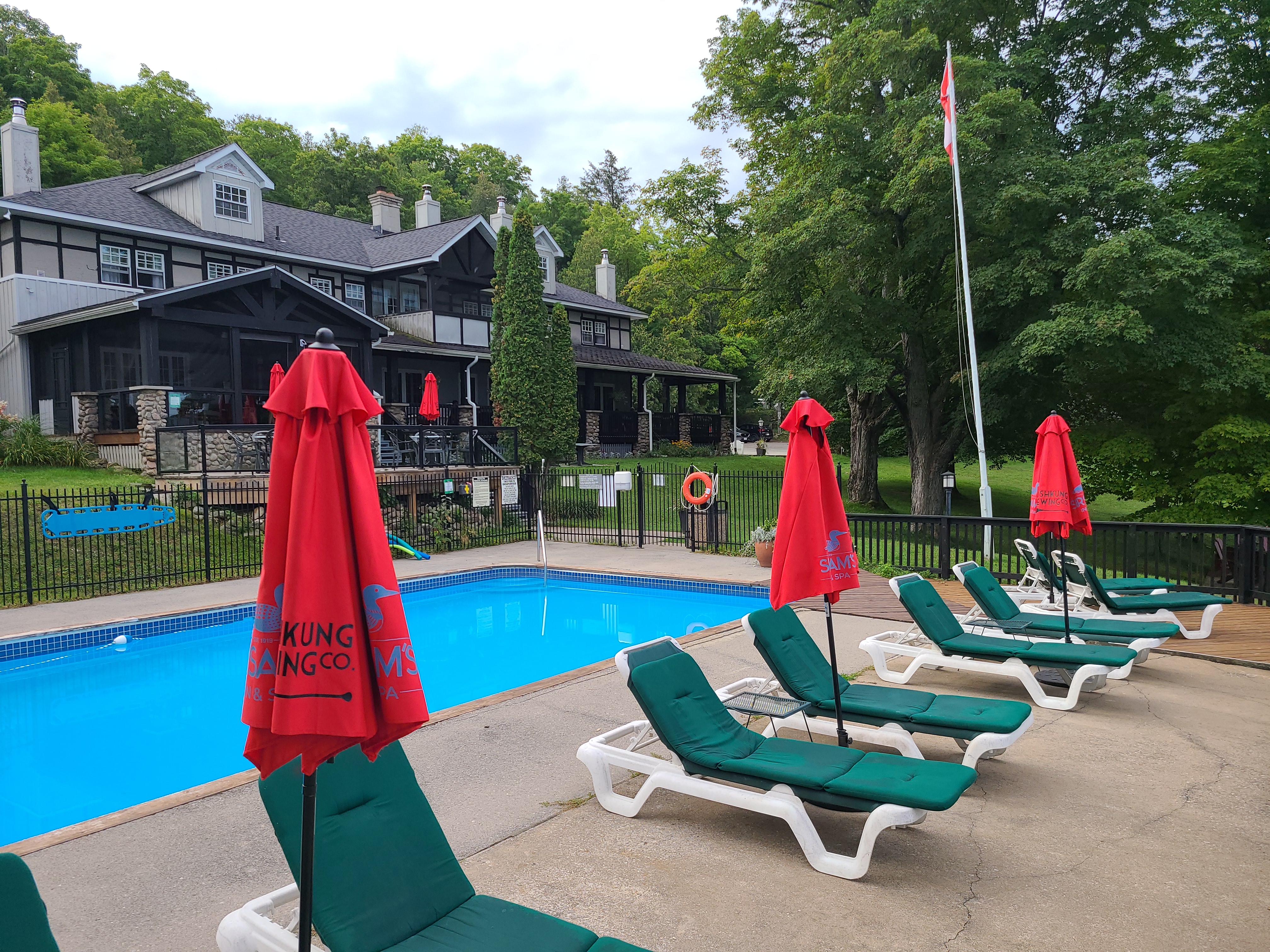white padded lounge chairs with red shade umbrellas line the deck of a luxurious outdoor swimming pool. In the background are lush green trees and a large stately resort lodge of dark brown wood.