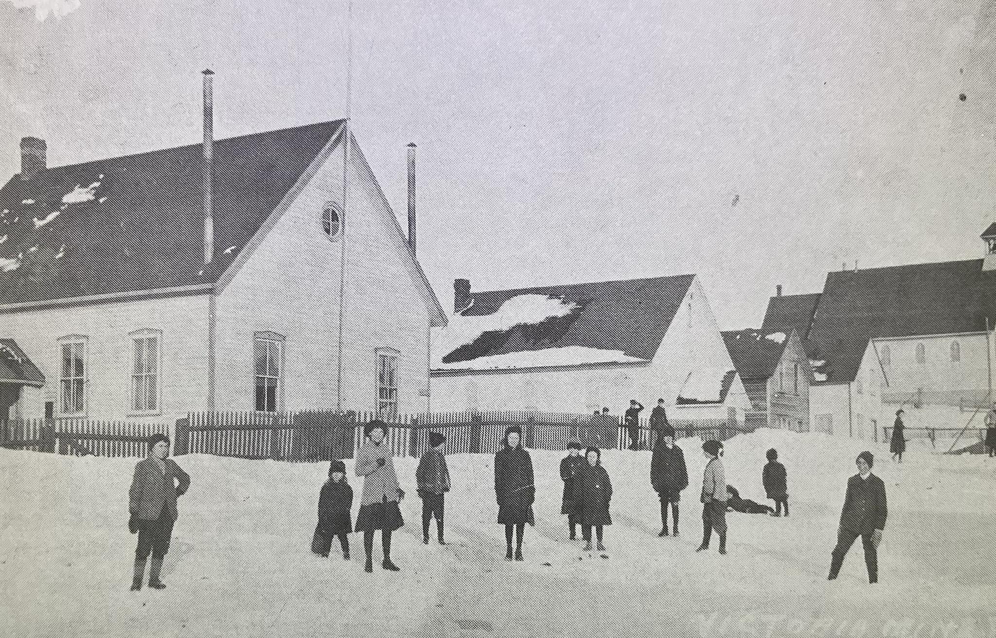A historic black and white photograph of a group of unidentified children standing in the snow outside the Victoria Mine schoolhouse that was moved to Coniston in the early 1900s.
