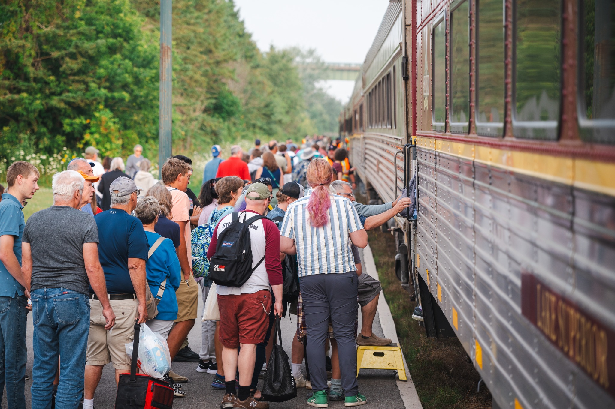 Many passengers gather next to the Agawa Tour Train to board. The train track is lined by thick green forest. 
