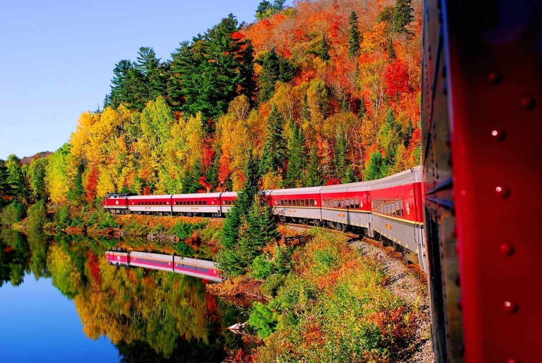 the red and silver Agawa Tour Train slides along the edge of a mirror-like lake reflecting both the train and the display of red, green yellow, orange and gold autumn leaves that fill the forested bank.