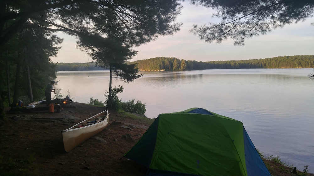 Tent, canoe and person standing by campfire on a campsite with lake in the background.