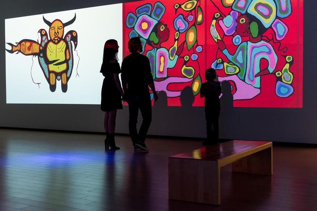 A family silhoutted in front of a wall of large illumintated paintings in vibrant colours at the Art Gallery of Algoma.