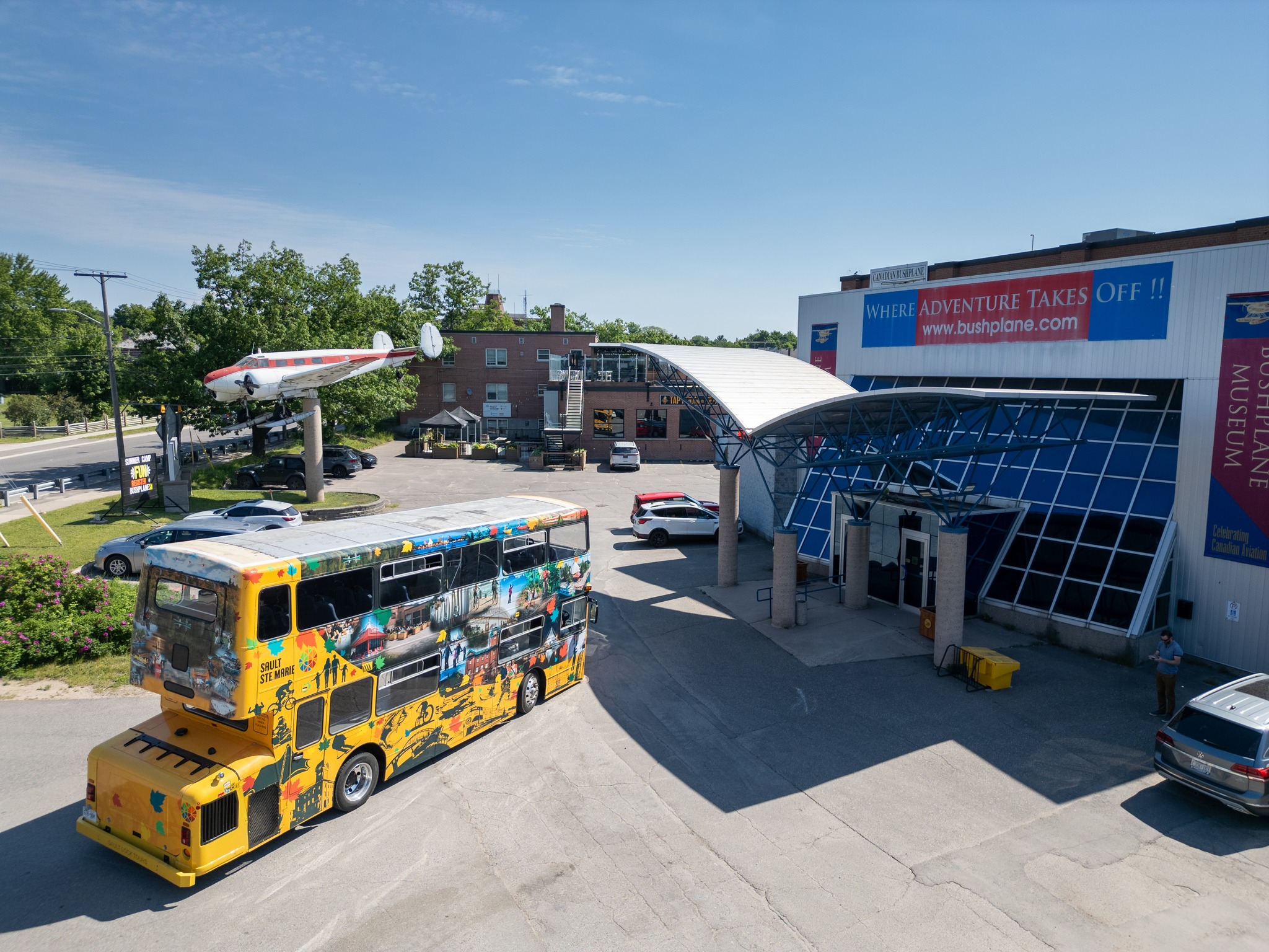 The yellow double-decker Big Ben Tour Bus sits outside the Canadian Bushplane Museum on a clear sunny summer day. The museum is large, modern, glass-fronted building with an antique bushplane displayed high on a pedistal outside.