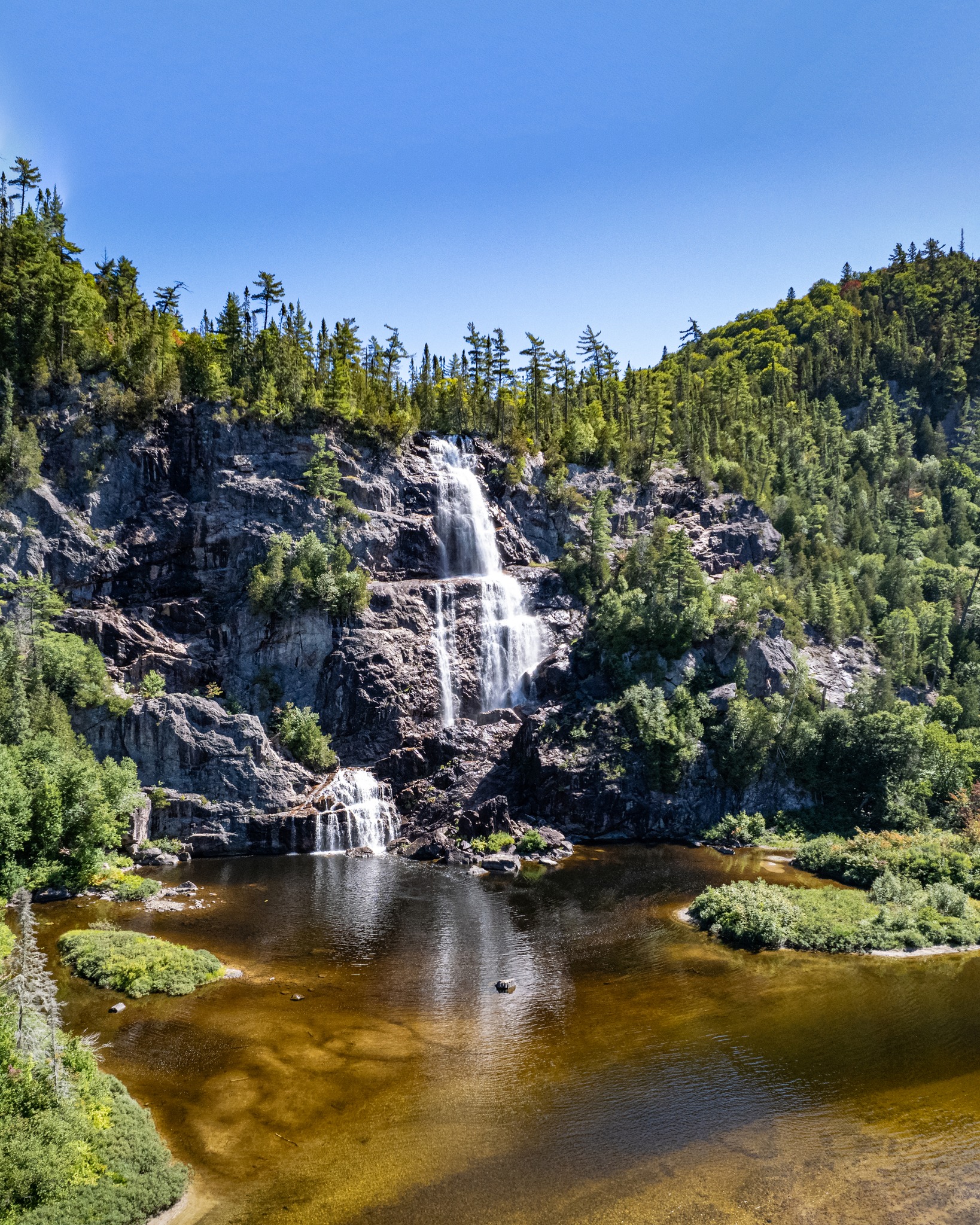 Bridal Veil Falls; a tall cascading falls rushes down a steep, forested rock face into a glassy pool under a clear blue summer sky. 