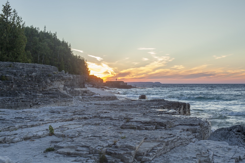 Person sitting at rocky shore with sun rising in background