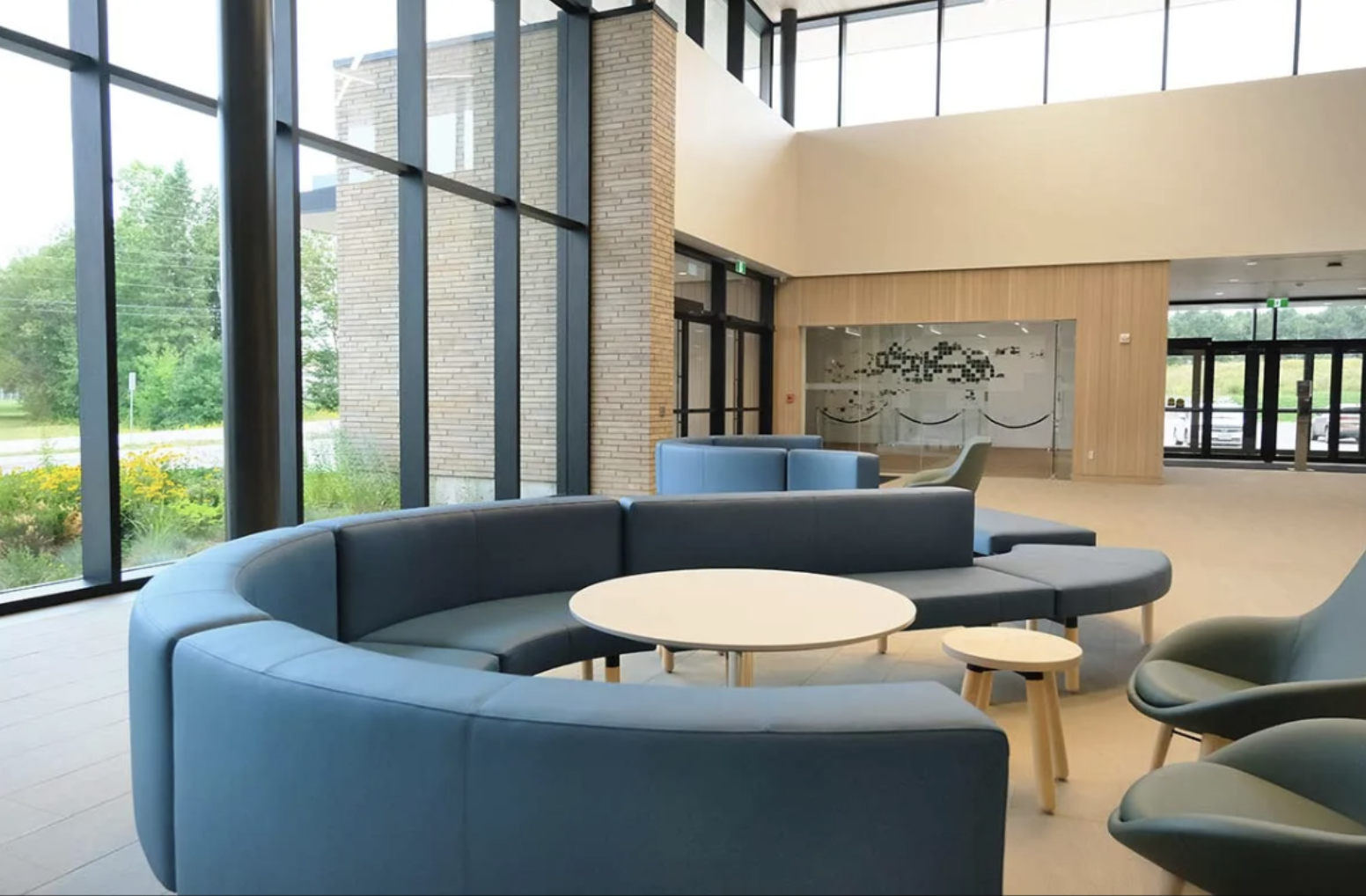 a sleek and modern meeting room with cozy circular blue couches and chairs around round tables, well-lit by a wall of tall windows.  