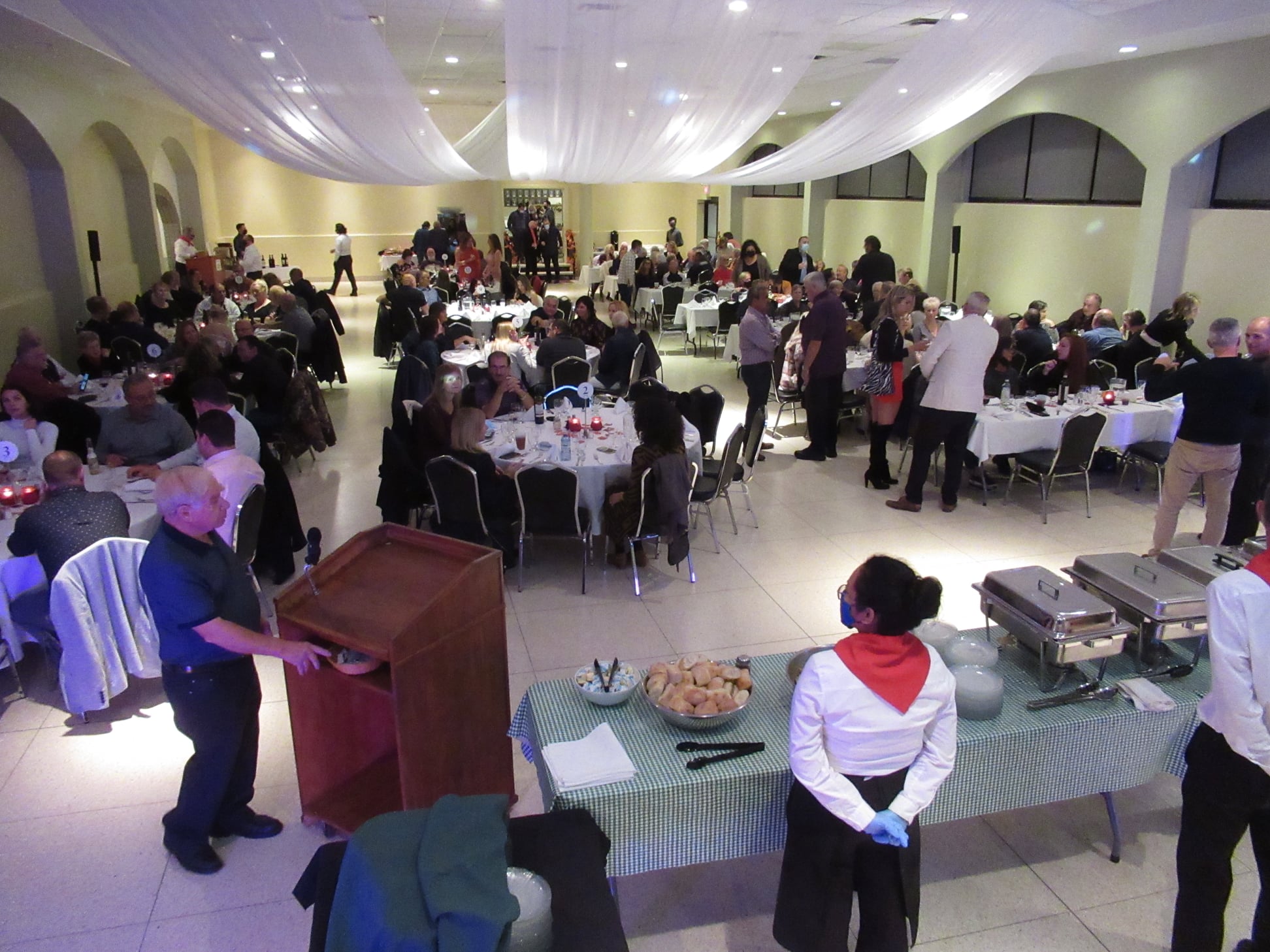 a crowd gathers around round tables in a banquet hall elegantly decorated with gauze drapings while banquet staff stand at attention around a table set for a buffet. 