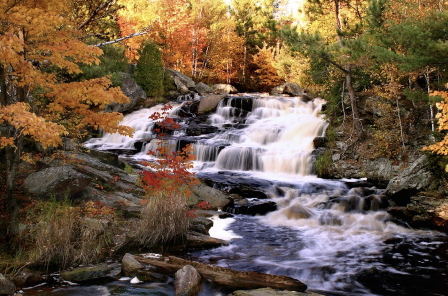 Duchesnay Falls, a gentle, cascading waterfall over rounded boulders surrounded by vibrant red, orange, yellow and green autumnal forest. 
