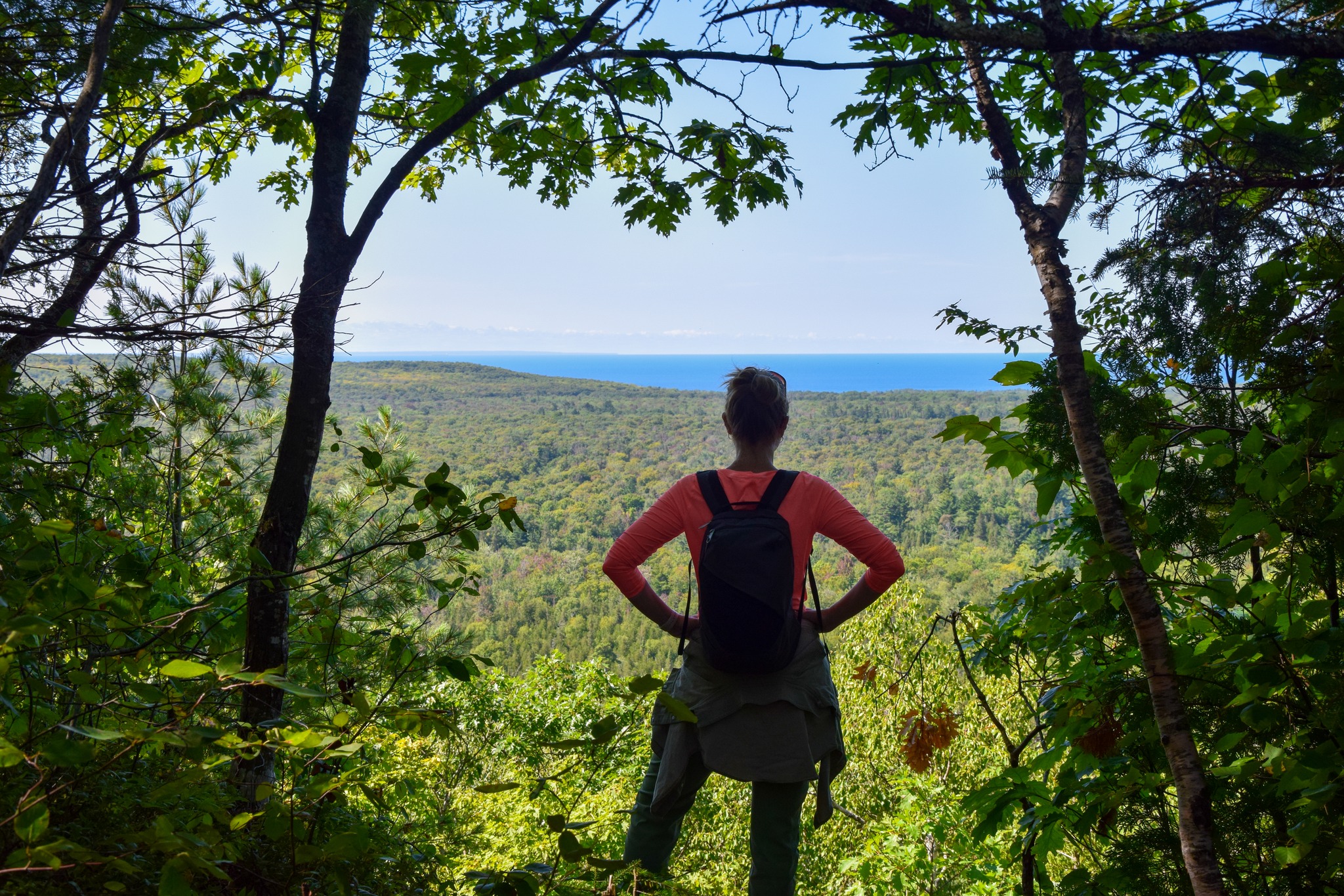 A hiker wearing a backpack stands with hands on hips, looking out through a clearing in the trees at a broad expanse of lush forest and blue lake to the horizon. 