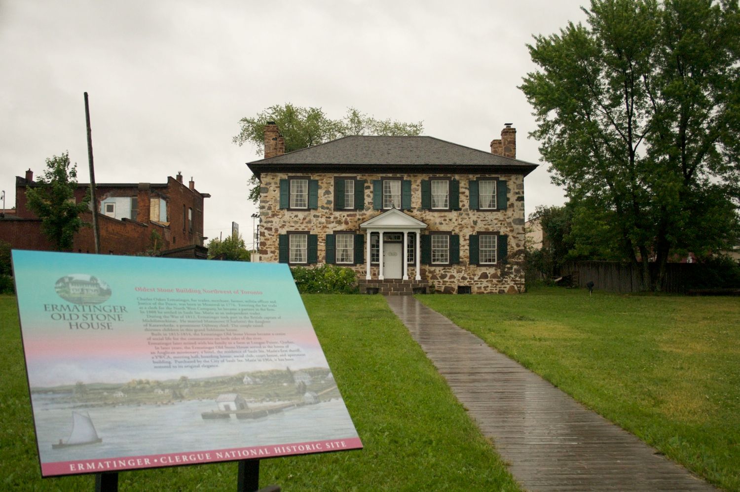 The Ermatinger Clergue Old Stone House: a stately stone mansion with rows of green shuttered windows and large old trees around it. An informational plaque sits at the end of a long walkway to its doors. 