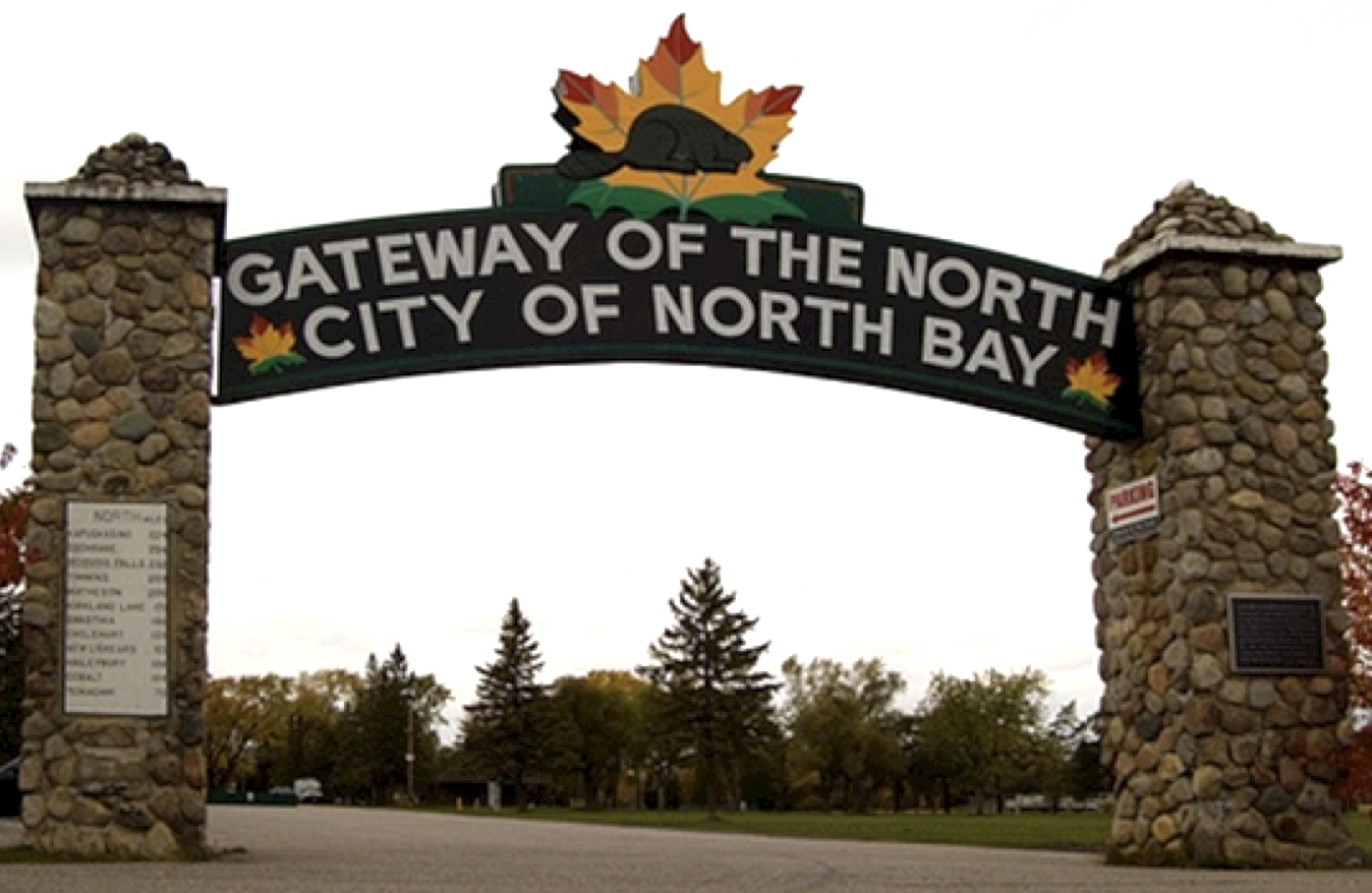 A stone archway with a sign reading "Gateway of the North, City of North Bay" on it, featuring an emblem of a maple leaf and a beaver. Green forest lies on the other side of the arch. 