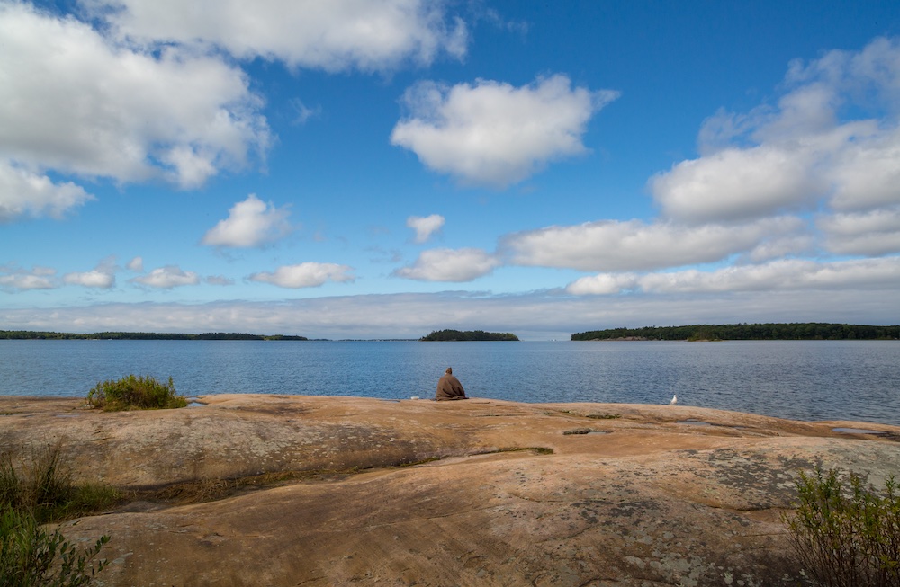 Person sitting at edge of rocky shore with big clouds overhead in blue sky