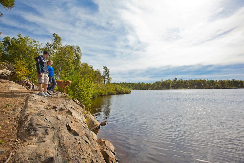 a man, a woman and a dog stand on a rocky bank looking happily out over a shining lake under a marbled blue sky. The leaves are starting to change for autumn with hints of orange and gold.  