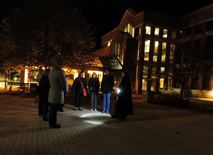 a group of people stand in the darkness next to the illumintated North Bay Courthouse while on a ghost tour.
