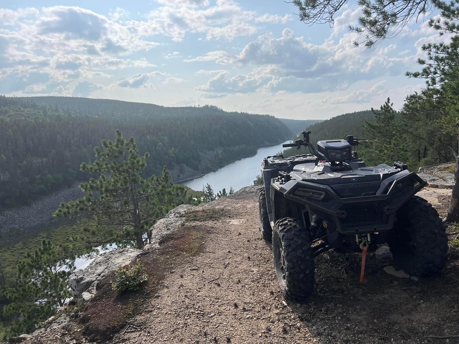 An ATV parked at the top of a cliff overlooking a thickly forested valley with a shining river at the bottom. 